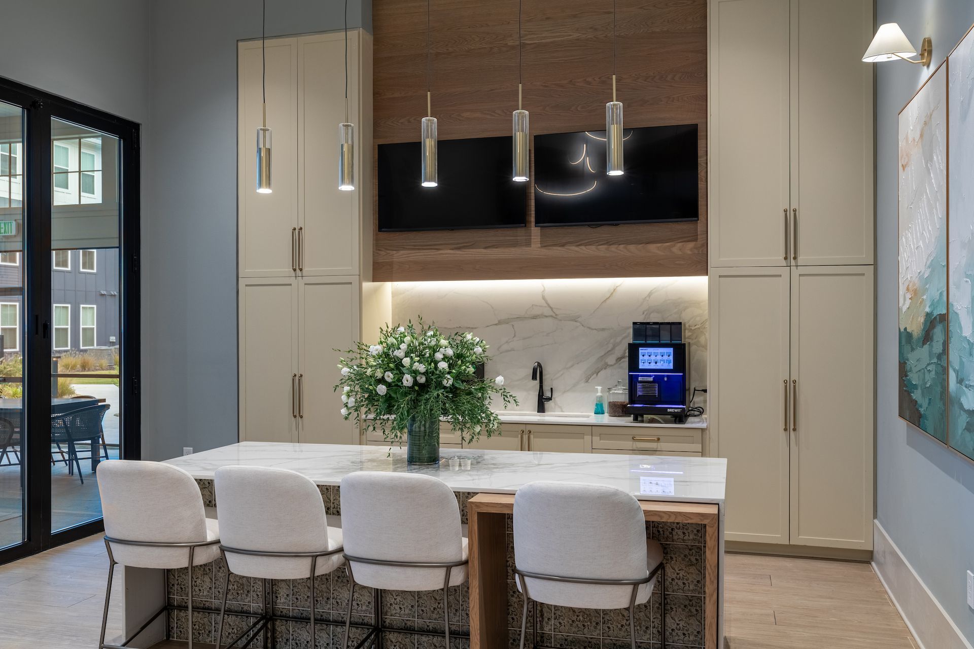Modern kitchen with island, bar stools, cabinetry, and marble countertops; a built-in coffee station and two TVs.