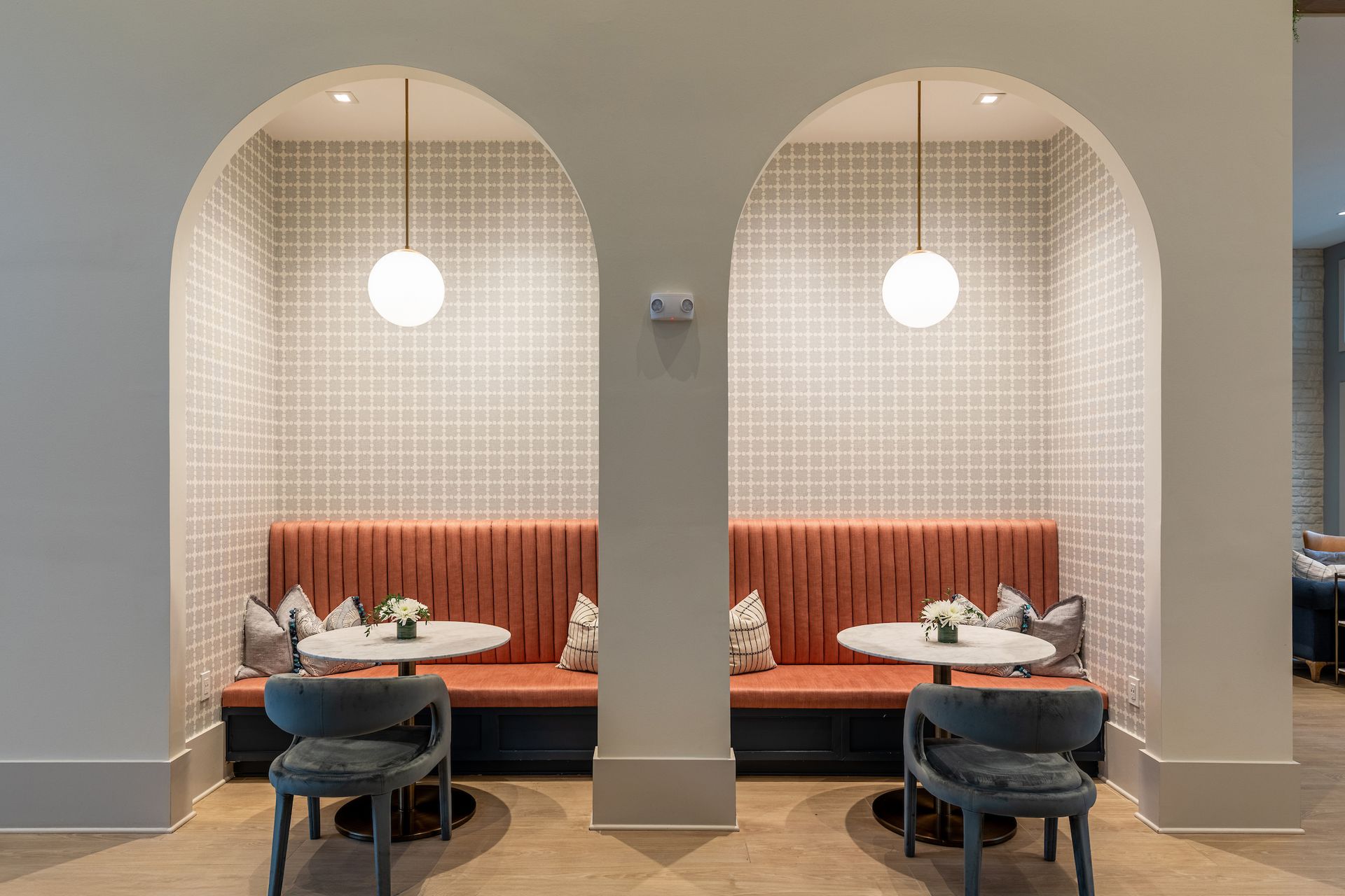 Two arched booths with coral benches, round tables, gray chairs, and patterned wallpaper, lit by globe pendants.