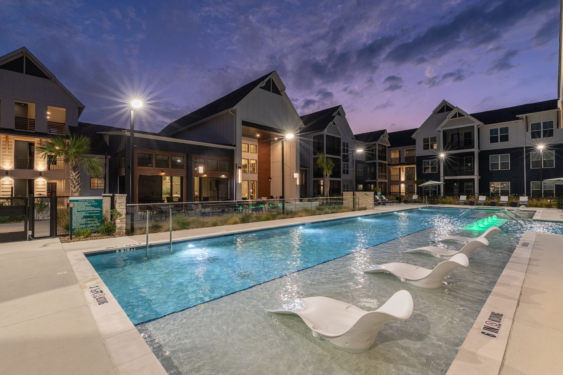 Night view of an apartment pool with in-water lounge chairs. Buildings surround the pool. Blue and teal hues.