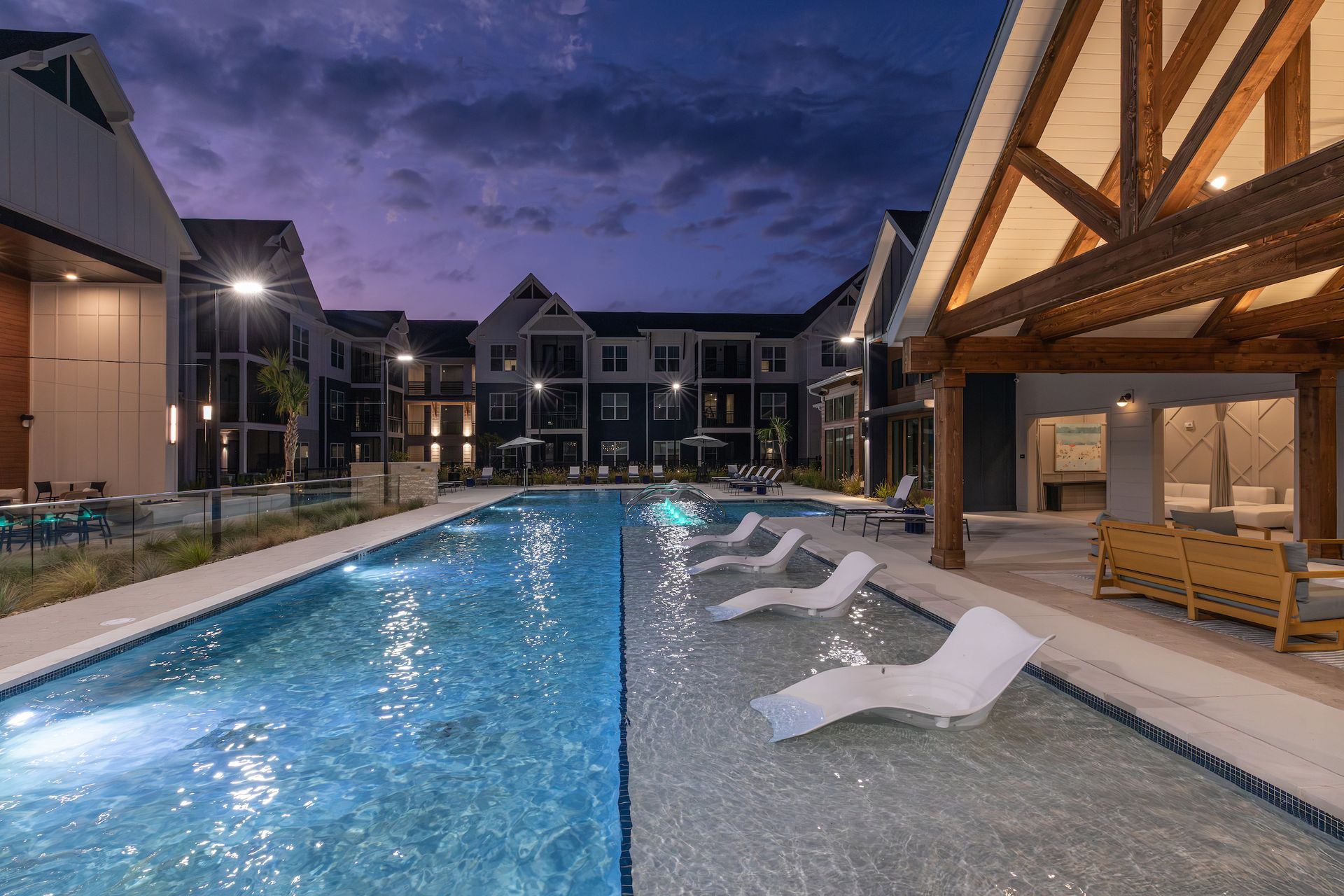 Nighttime pool with lounge chairs and apartment buildings under a twilight sky.