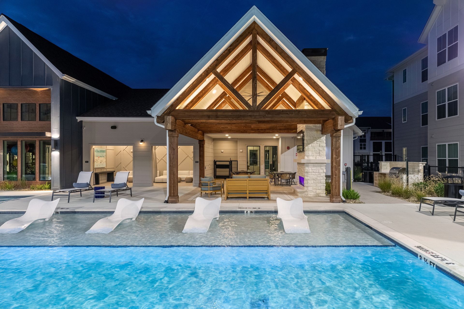 Pool area at night, with white lounge chairs in the pool, covered patio, and apartment building in the background.