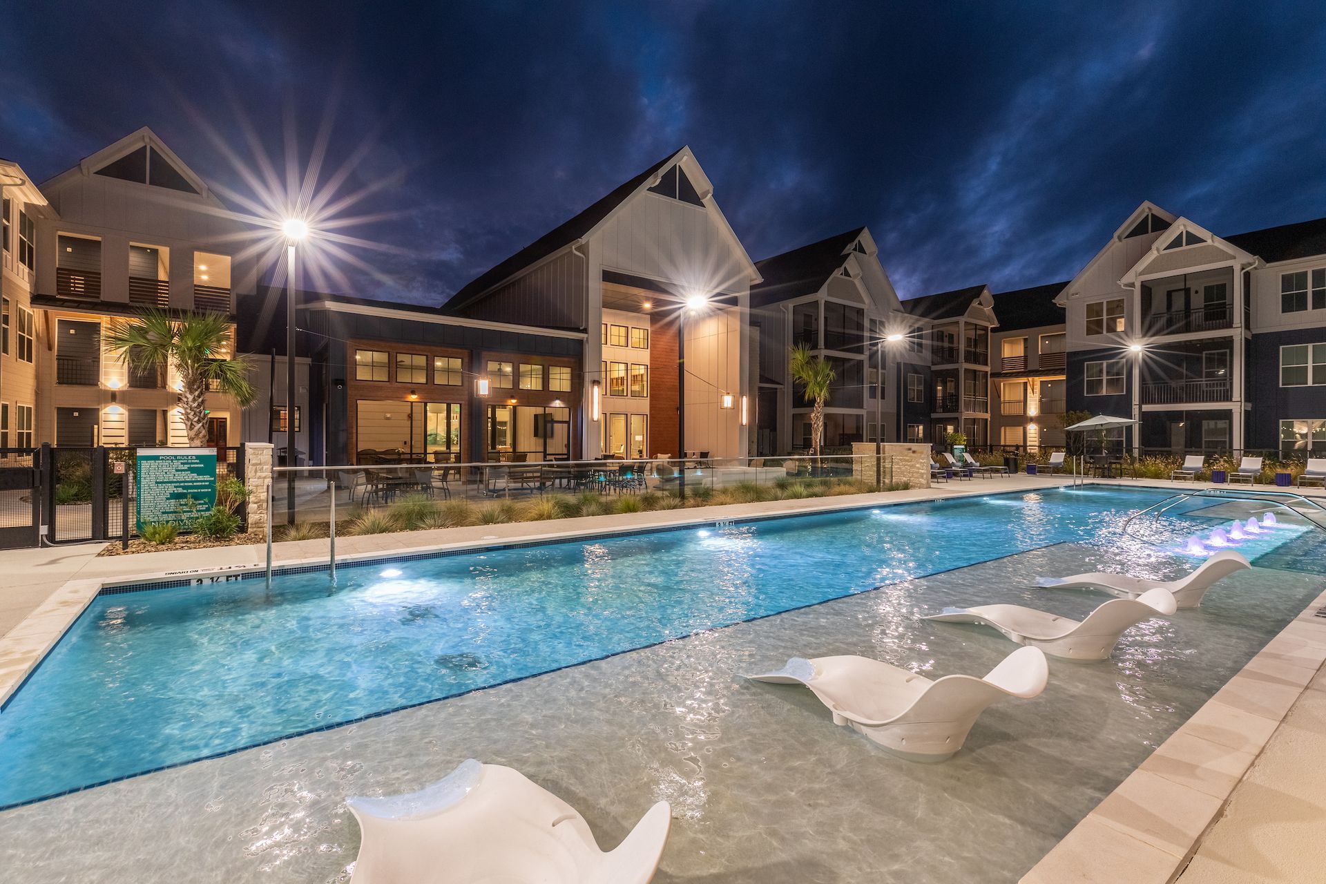 Nighttime view of an apartment complex with a pool. White lounge chairs in shallow water, buildings with bright lights.