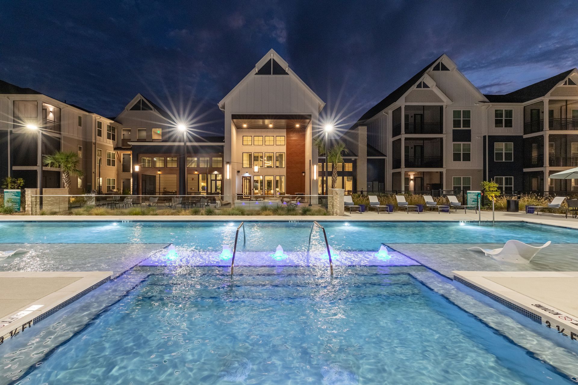 Nighttime view of a luxury apartment complex with a lit swimming pool and fountains.