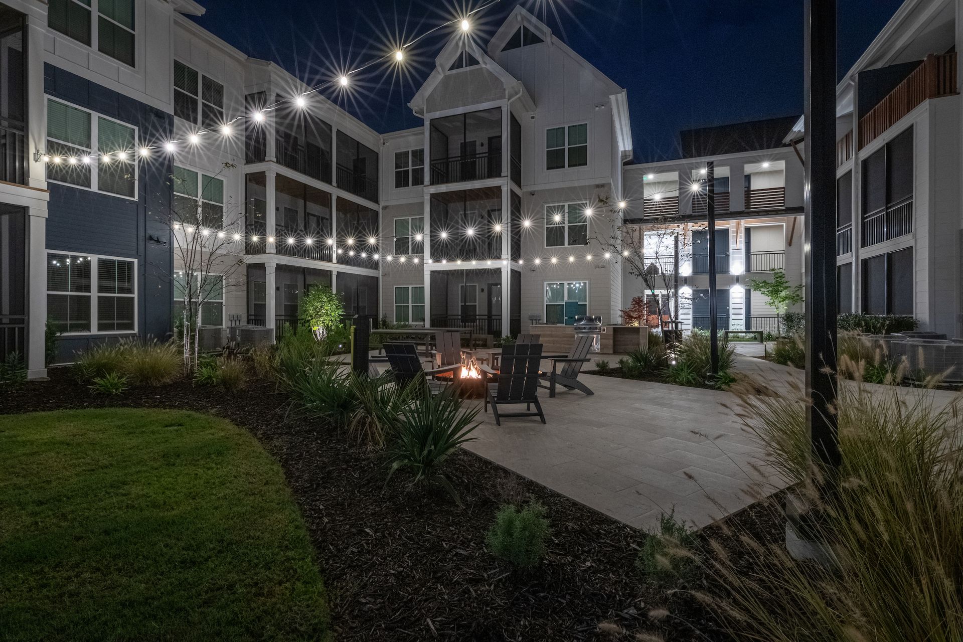 Night view of apartment courtyard with string lights, fire pit, and seating.