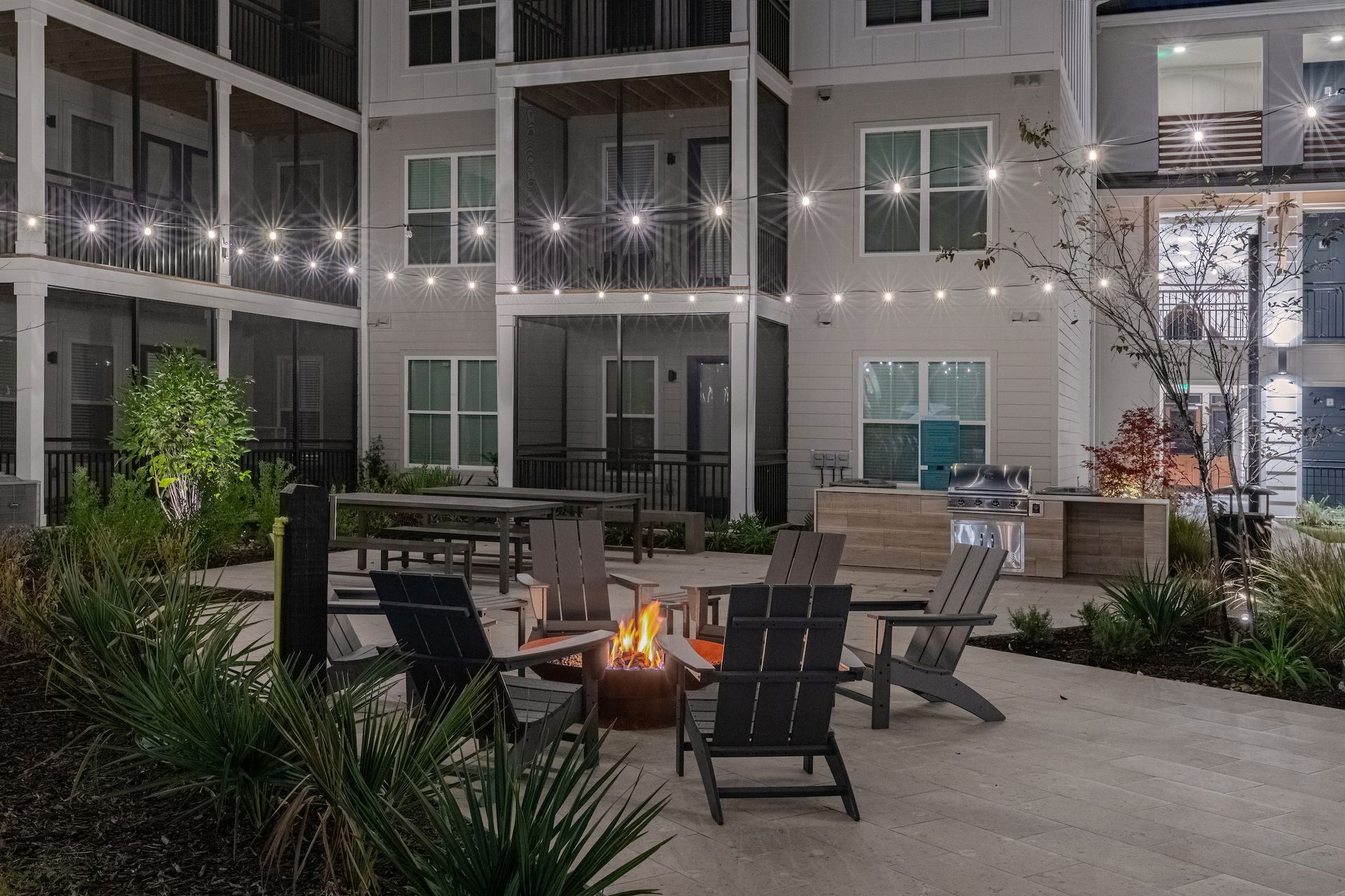 Courtyard with fire pit, seating, string lights, and grill in front of a modern apartment building at night.