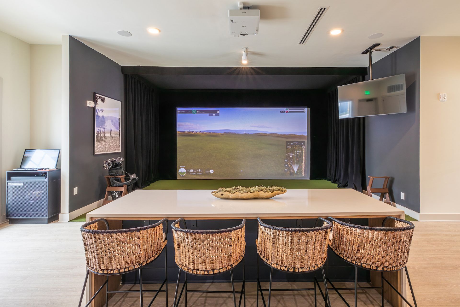 A room with a golf simulator. A large screen displays a golf course, and a bar with chairs is in front.