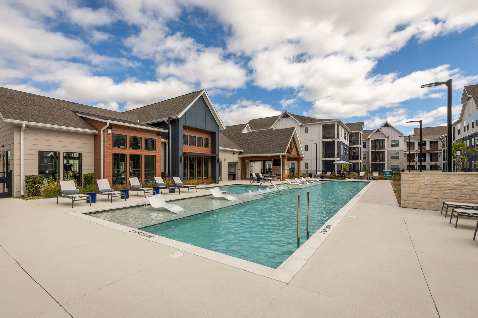 Pool and apartment complex with blue water, white lounge chairs, and buildings with blue and brown exteriors.