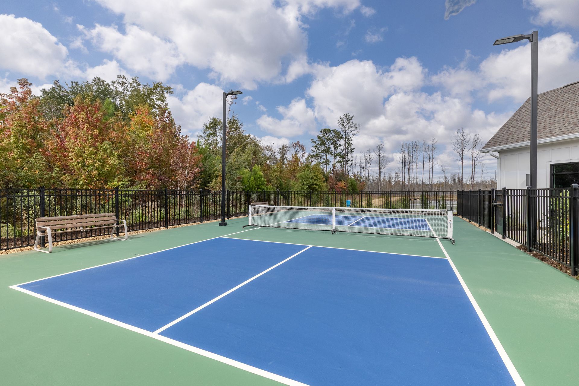 Pickleball court with blue playing surface, green surround, net, fence, bench, and cloudy sky.