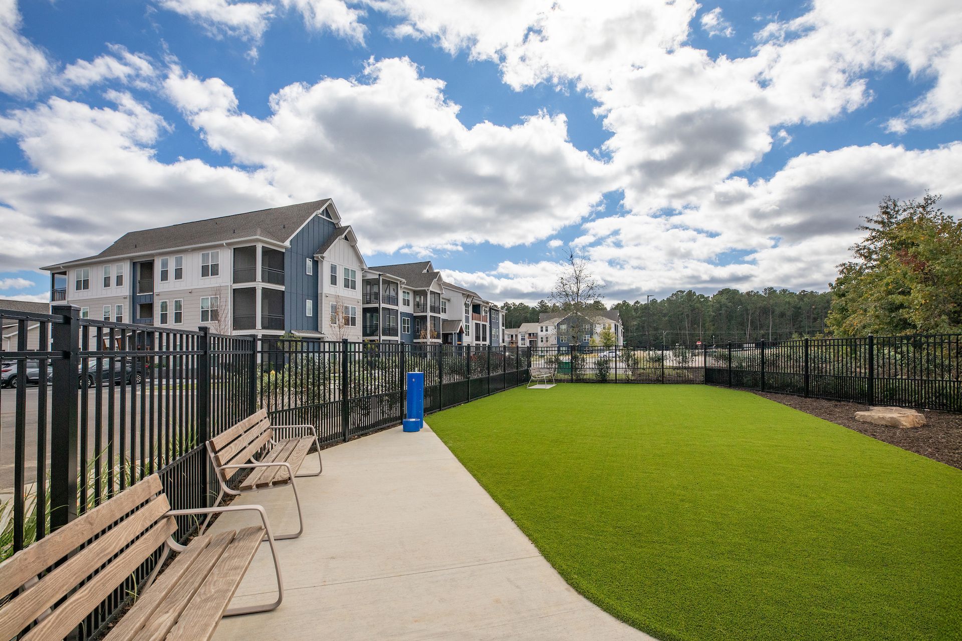 Dog park with benches, artificial turf, and fenced in, next to apartment building on a sunny day.