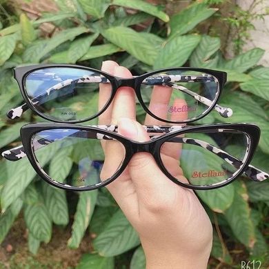 Una mano sostiene dos monturas de gafas negras con lentes espejadas sobre hojas verdes.