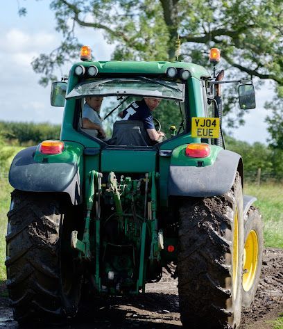 A green tractor driven through a field with two people visible in the cab, seen from the rear on a dirt track.