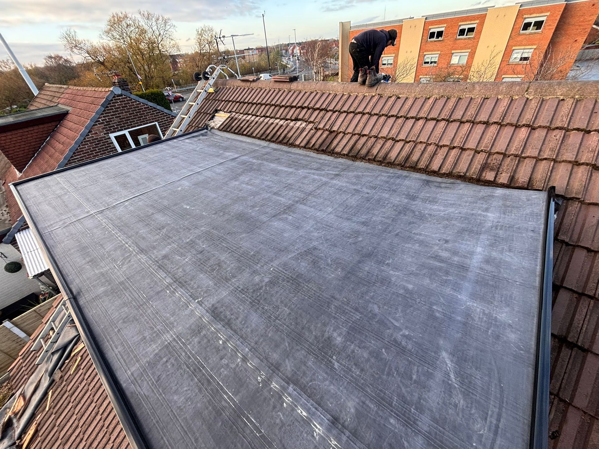 A person working on a flat, dark grey roof membrane surrounded by sloped, terracotta-tiled roofs.