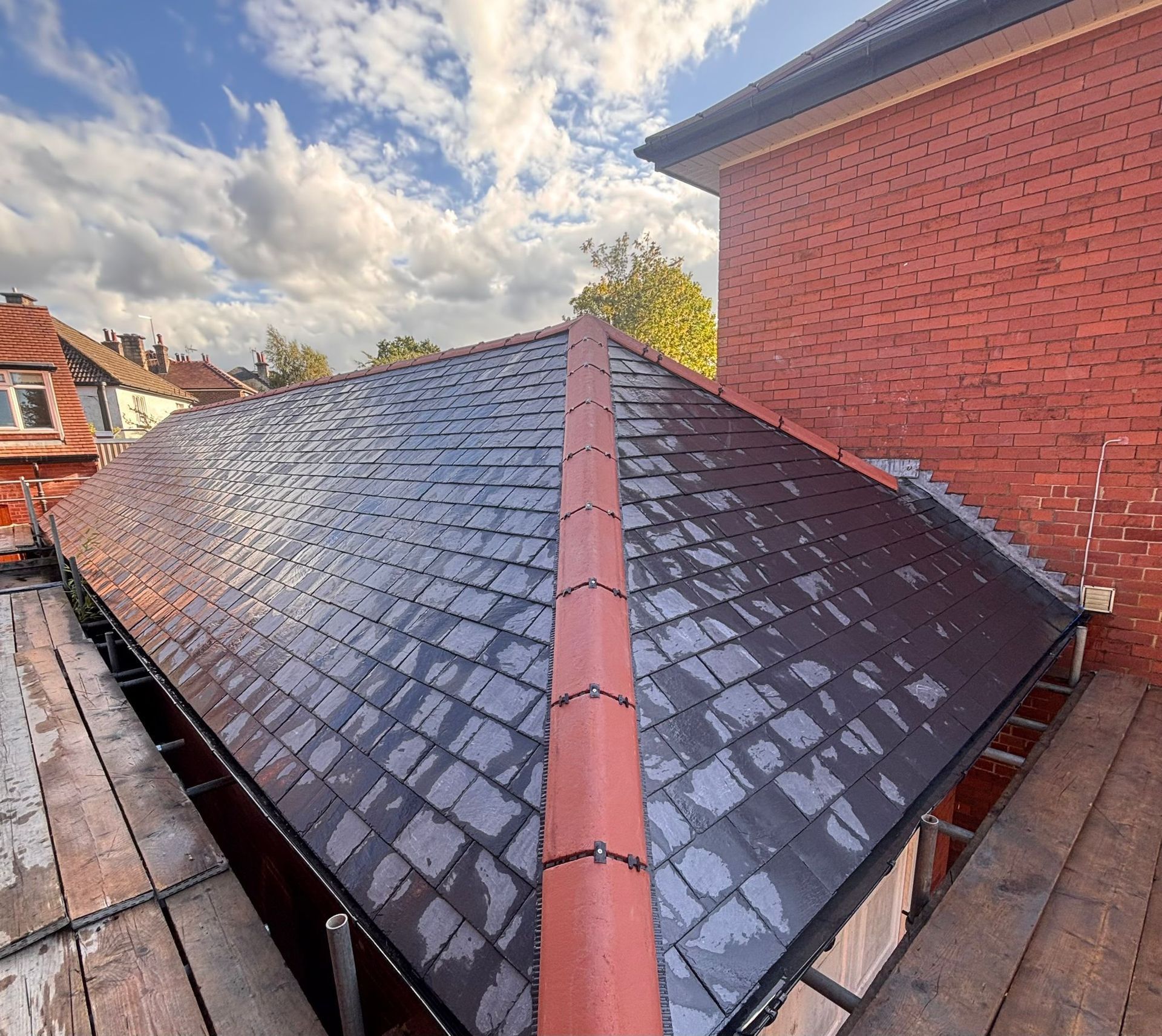 A newly tiled dark gray roof with red ridge tiles, seen from scaffolding under a blue sky with white clouds.