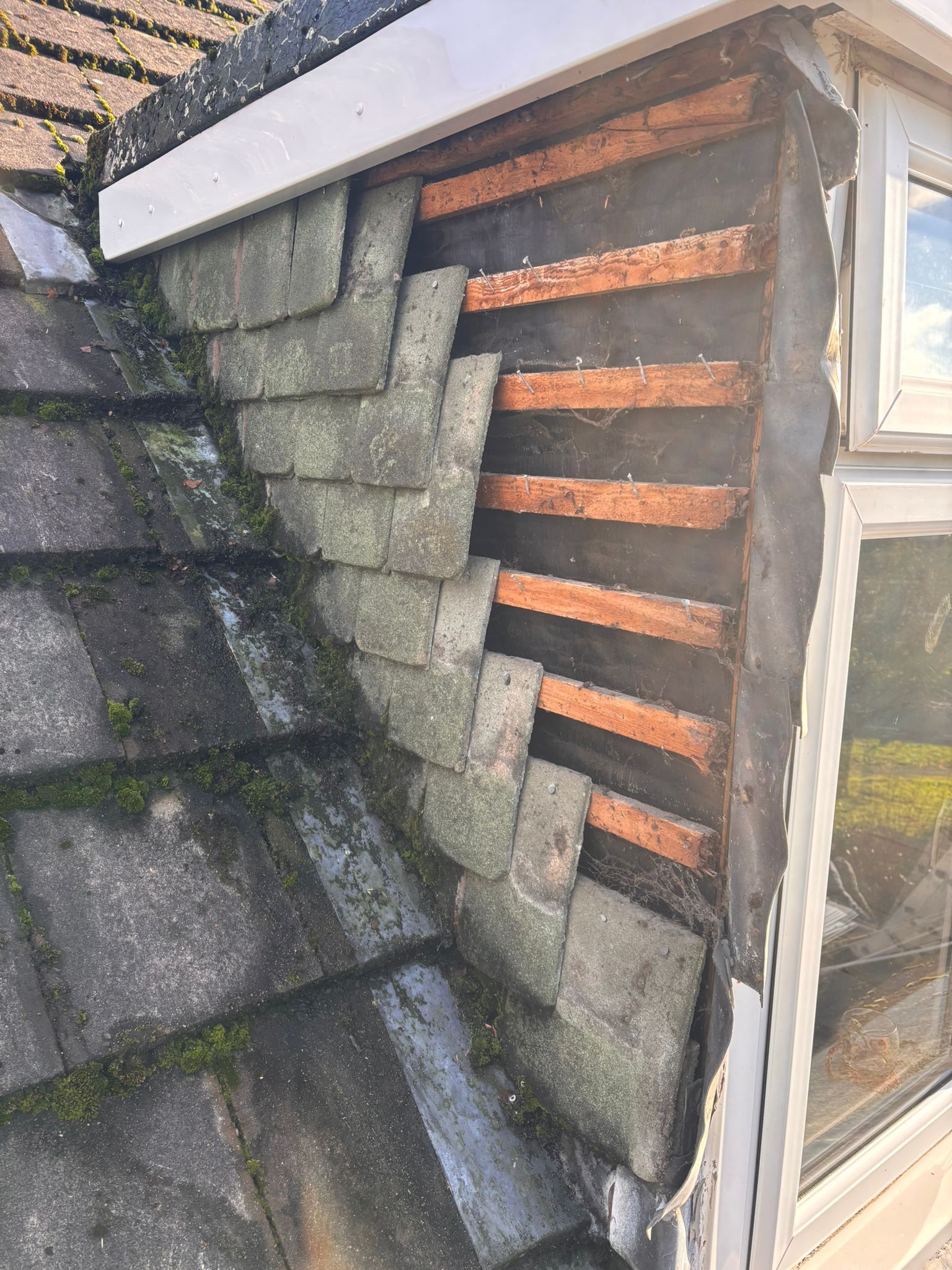 A damaged section of a pitched roof showing missing grey slate tiles, exposing horizontal wooden battens and dark felt.