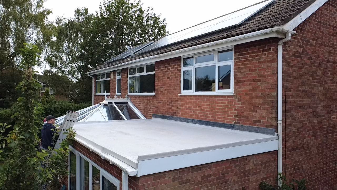 A person stands on a ladder working on the roof of a single-story brick extension attached to a two-story brick house.