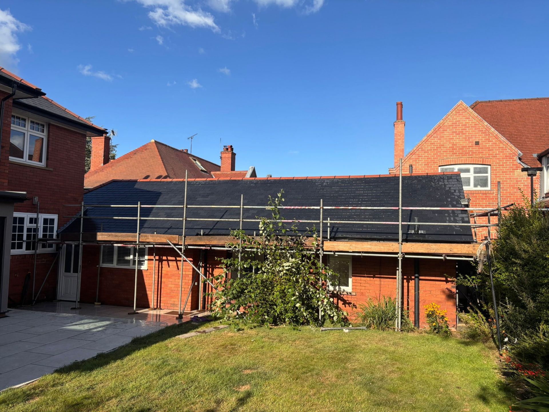 A single-story brick building under renovation with a black roof membrane and metal scaffolding, viewed from a garden.
