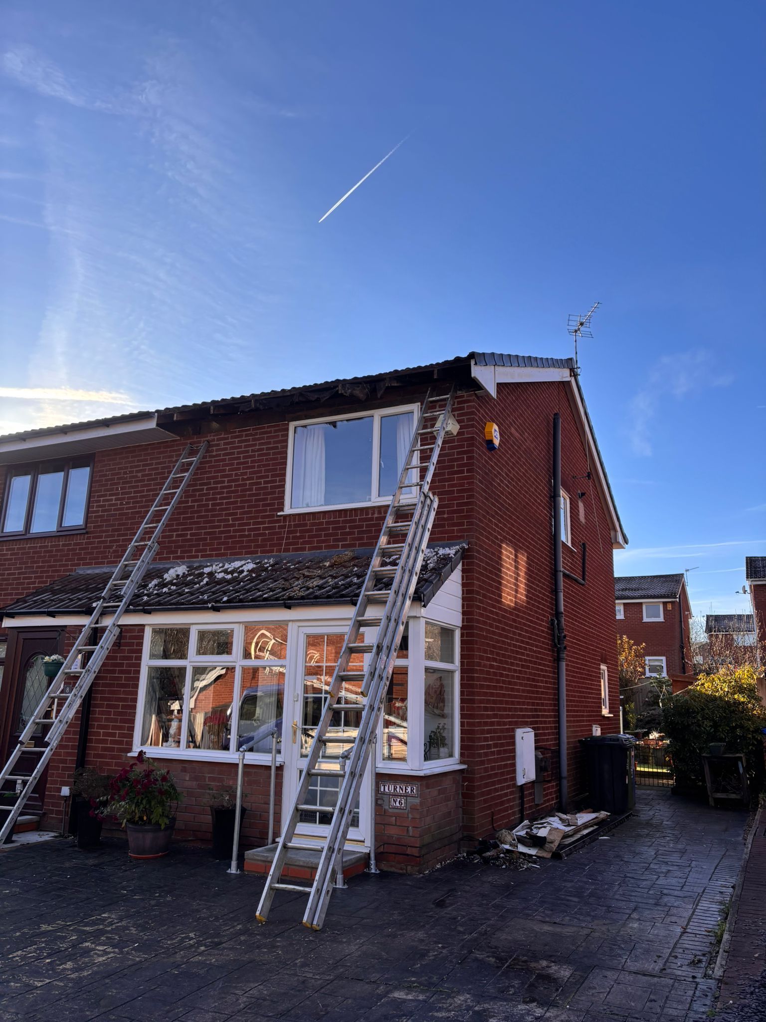 Two tall extension ladders lean against the front of a red brick house under a bright blue sky.