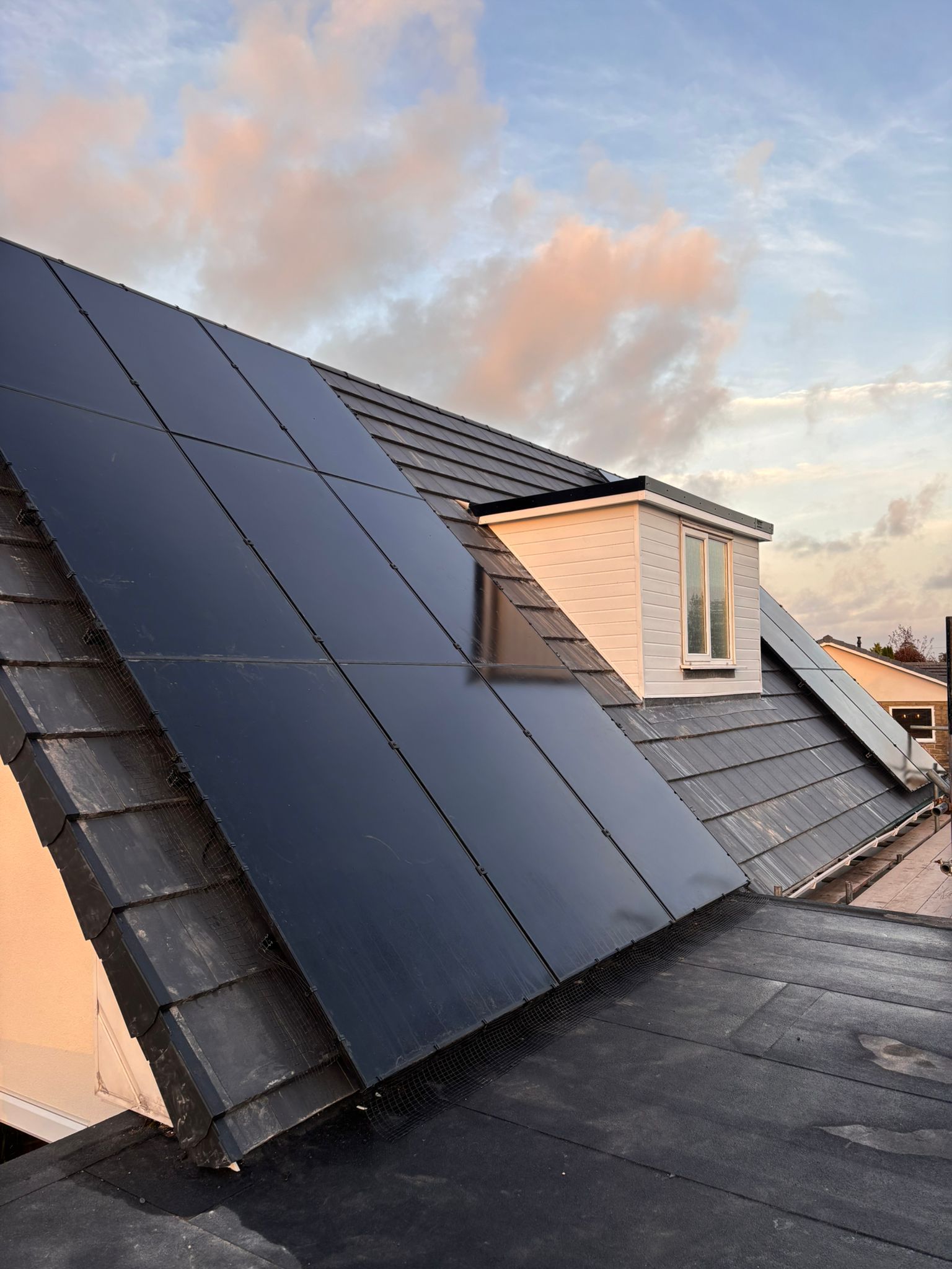 Black solar panels installed on a residential dark-tiled roof next to a white dormer window under a cloudy sunset sky.