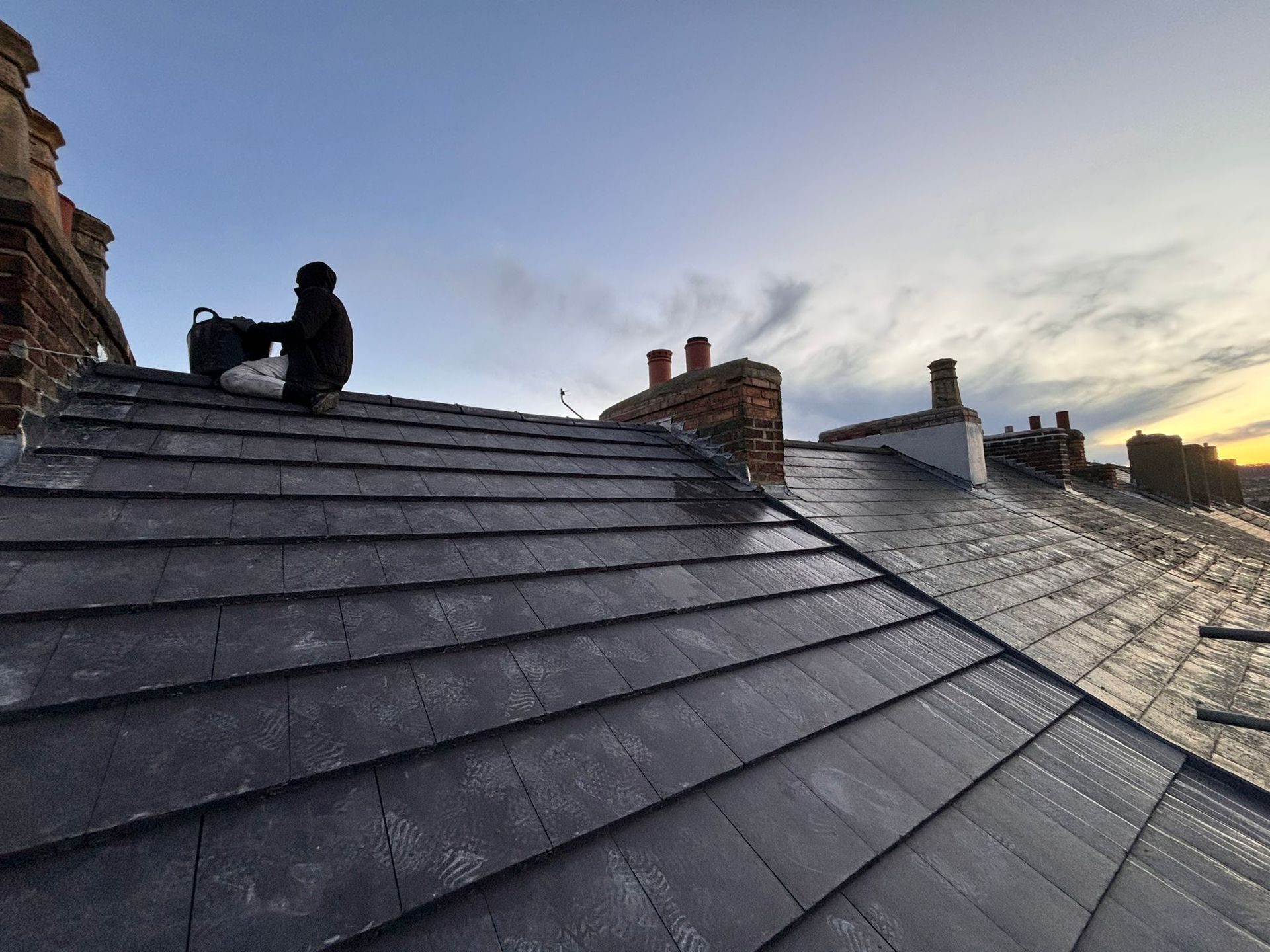 A person sits on a dark, slate-tiled roof at sunset, with multiple brick chimneys visible against a cloudy sky.