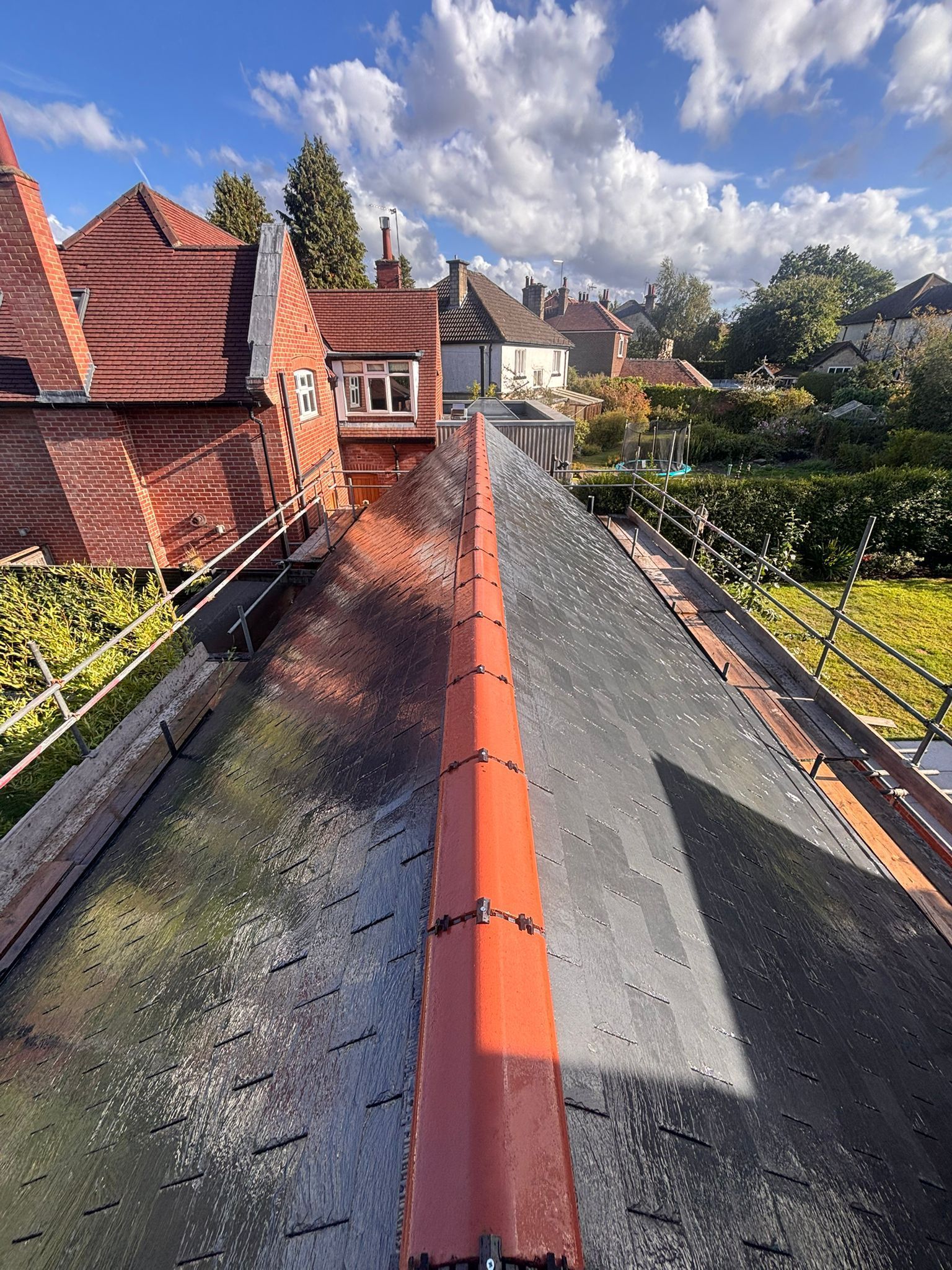 A high-angle view of a residential roof ridge featuring red tiles against a bright blue sky with scattered clouds.