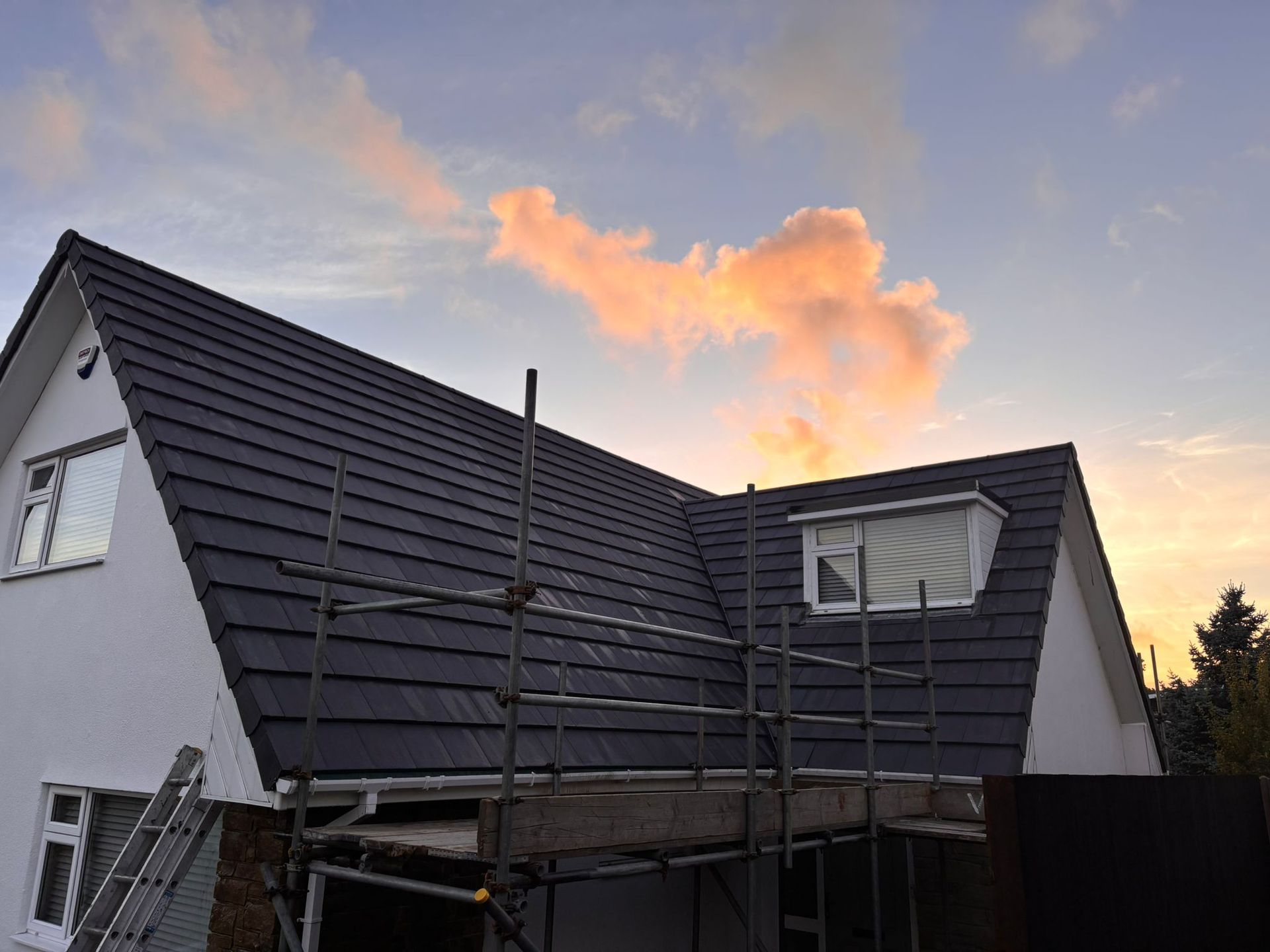 A house with a dark gray tiled roof and white walls under a sunset sky, with scaffolding set up along the side.