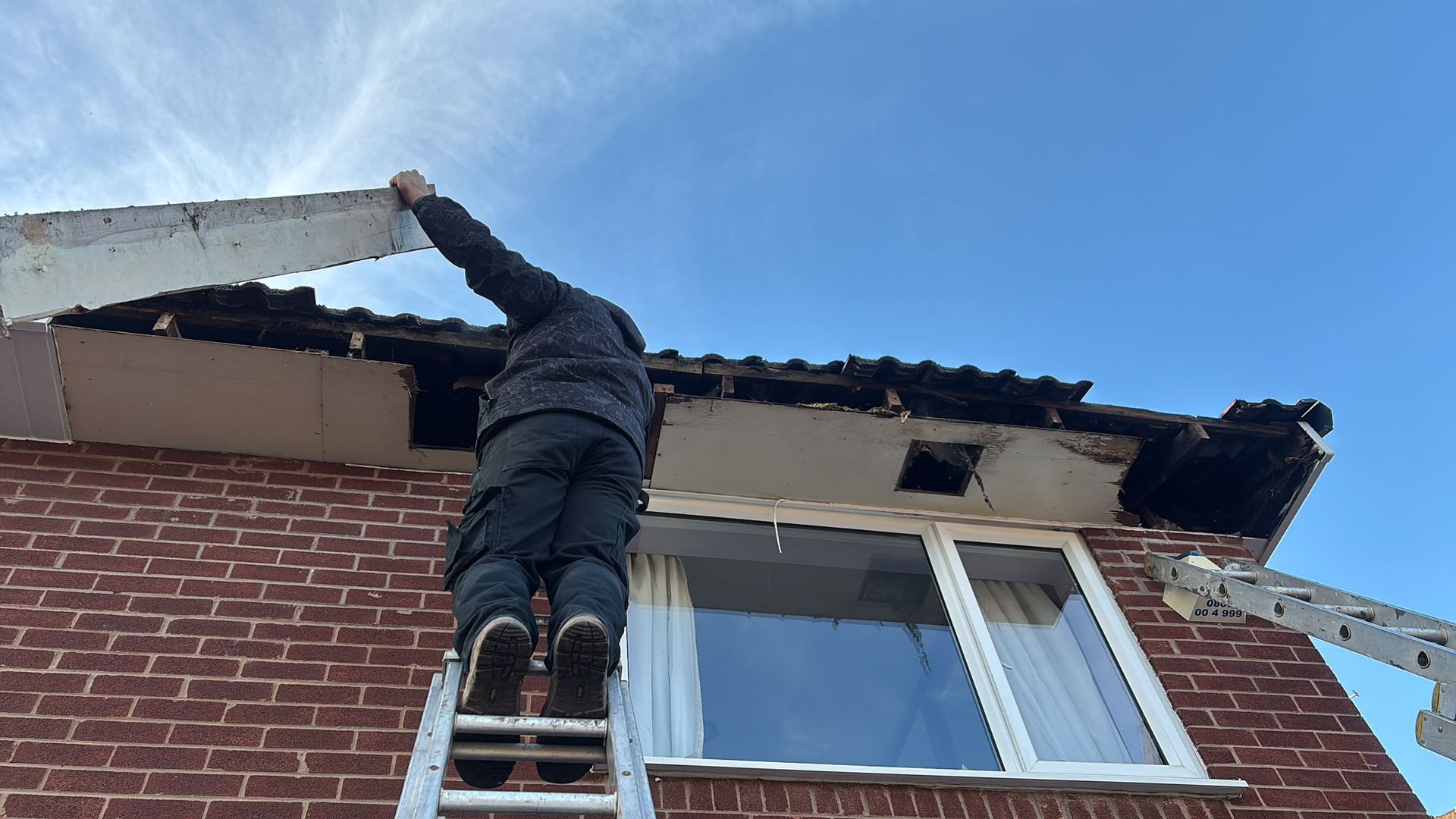 A person on a ladder removes a section of the white soffit from under the roof eaves of a brick house.