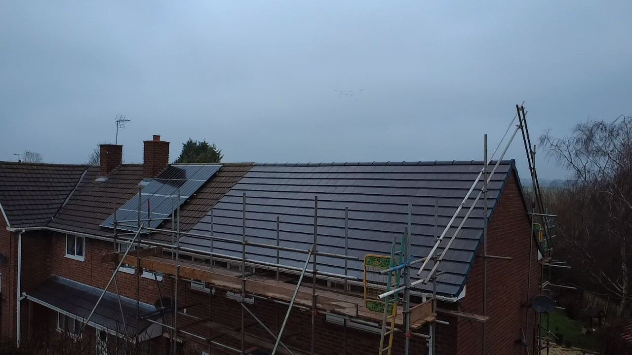 A residential brick house with a partially tiled roof, surrounded by scaffolding and featuring installed solar panels.