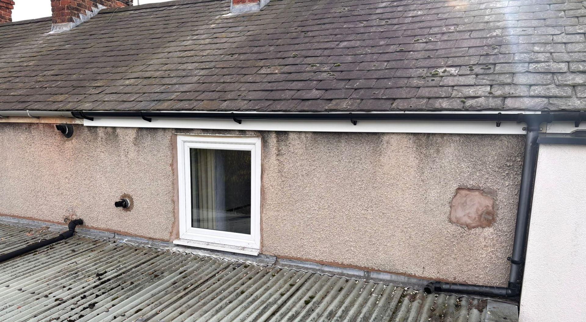 Exterior wall with a window, corrugated metal roofing, and a tiled roof above against a light-colored pebbledash surface.