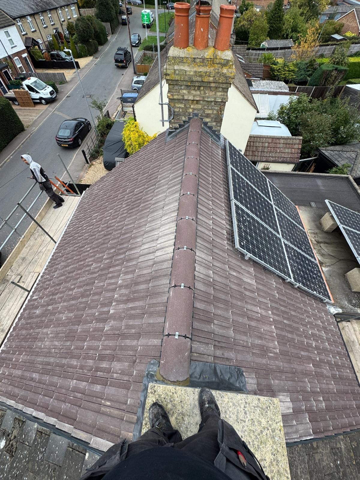 A first-person view looking down a tiled pitched roof toward a brick chimney with three pots, next to solar panels.