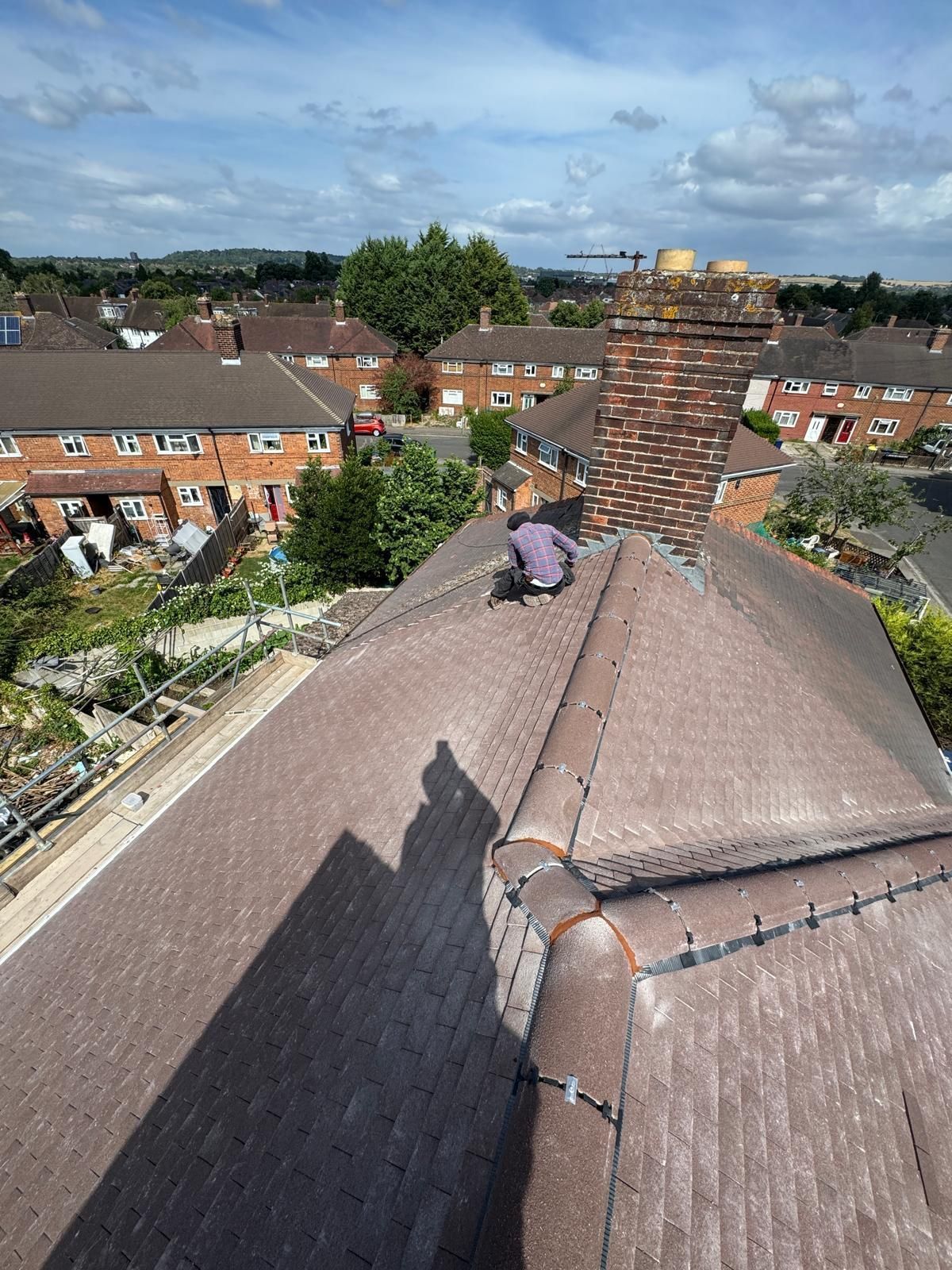 A high-angle view of a worker on a tiled residential roof performing repairs next to a large brick chimney.