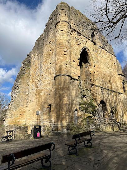 The stone ruins of Tonbridge Castle tower over a paved courtyard with benches under a partly cloudy blue sky.