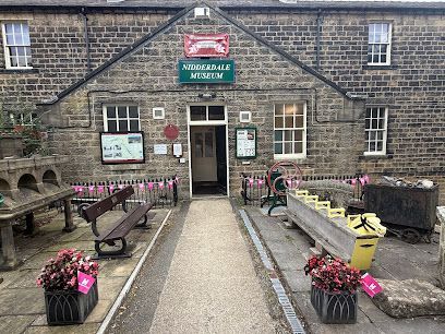 The stone facade of Suddersale Museum with a central doorway, benches, and outdoor artifacts on a paved path.