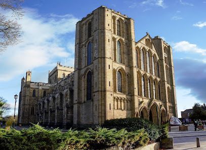 A low-angle view of the large, tan stone Ripon Cathedral with its tall twin towers under a bright blue sky.