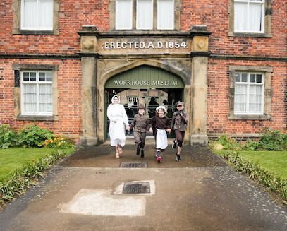 Four people dressed in historical clothing walk out of the arched entrance of the brick Workhouse Museum building.