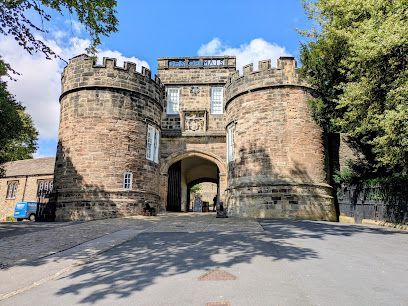 Stone gatehouse with twin towers and crenelated roof, flanked by trees and a paved path, under a blue sky.
