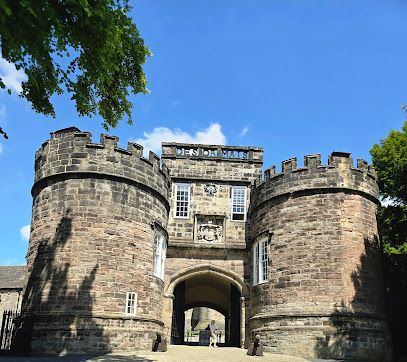 A stone gatehouse with two round towers, crenellated walls, and a central archway against a bright blue sky.