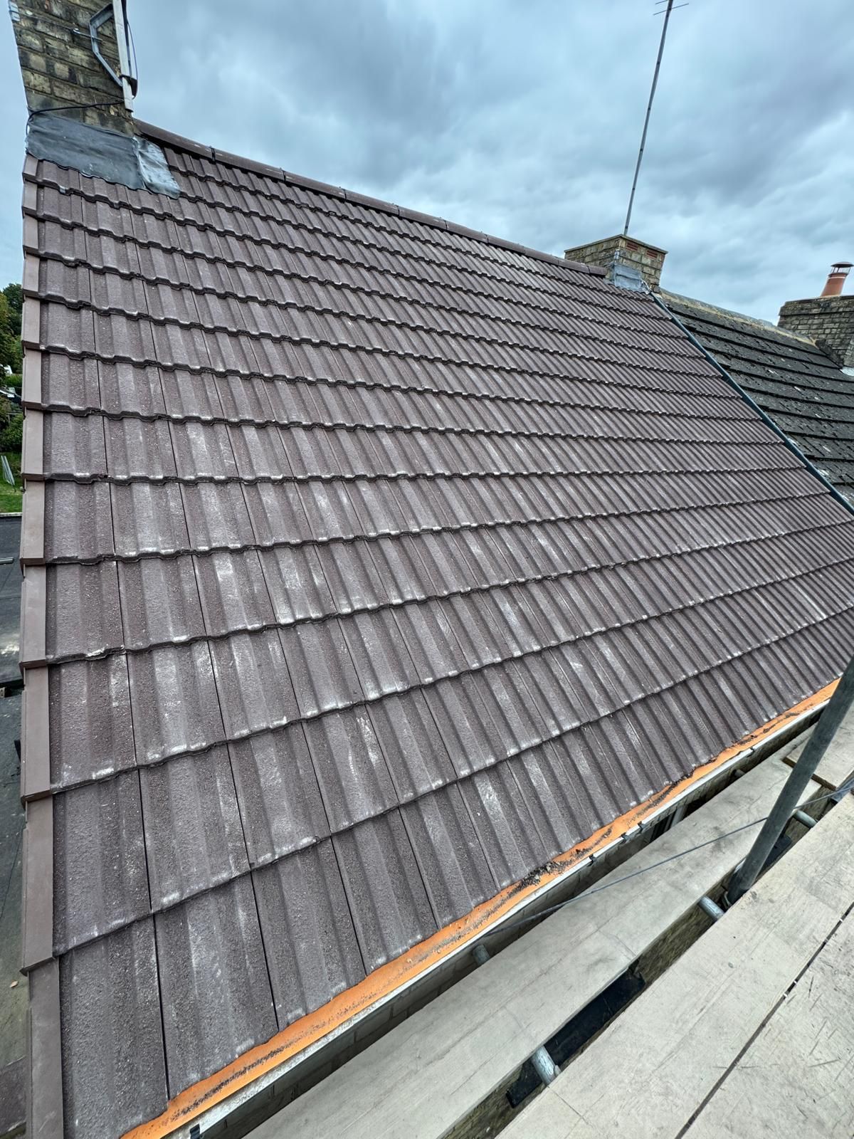 A steep, brown-tiled residential roof viewed from scaffolding against a cloudy sky.