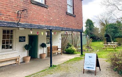 A brick building with a white storefront, a wooden bench, two potted trees, and an A-frame sign pointing to a gallery.