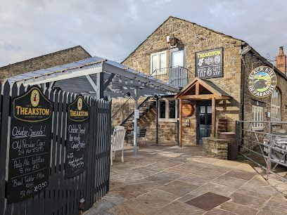 A stone building with a Theakston pub sign, an outdoor seating area under a pergola, and chalkboards with drink prices.