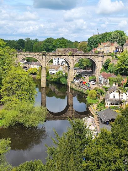 A stone arched railway bridge spanning a river, surrounded by lush green trees and a small town on a sunny day.