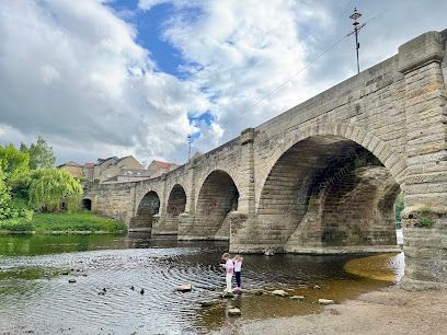 Two children standing in a shallow river beneath a large, historic stone arch bridge under a cloudy sky.