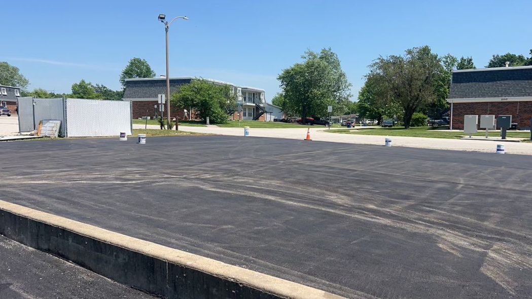 A freshly paved black asphalt parking lot under a clear blue sky, bordered by a concrete curb and surrounding buildings.