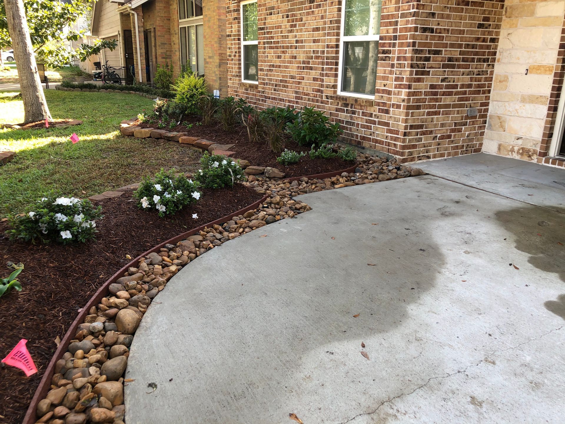 A curved driveway next to a garden bed filled with plants, mulch, and rocks, adjacent to a brick house.