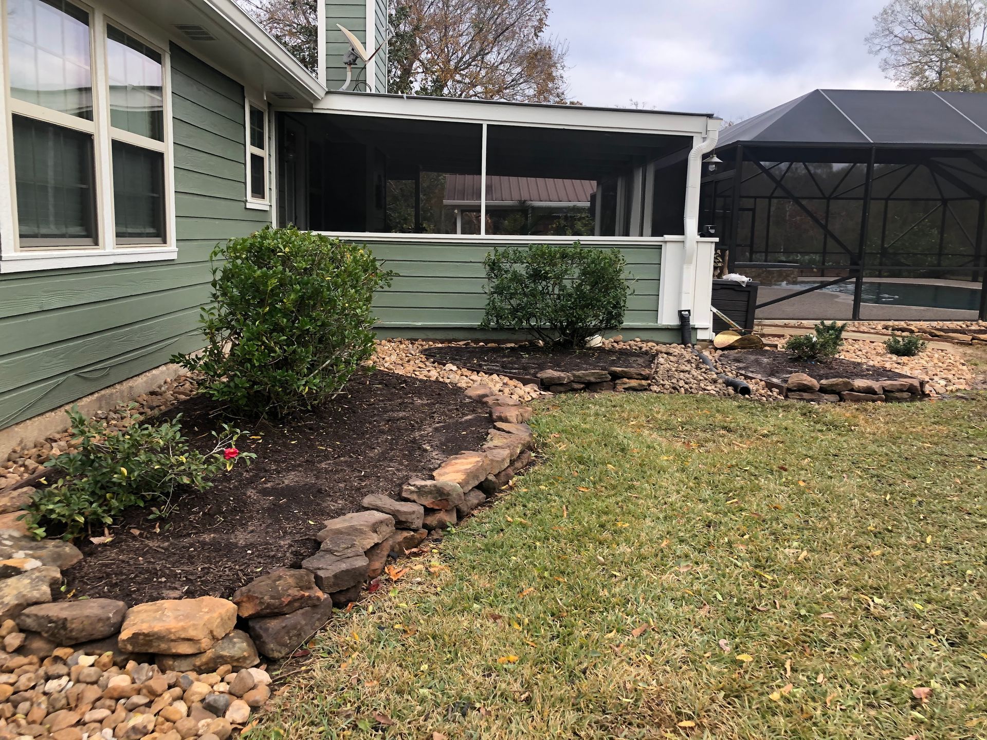 Green house with flower beds bordered by rocks, with screened porch and pool enclosure in background.