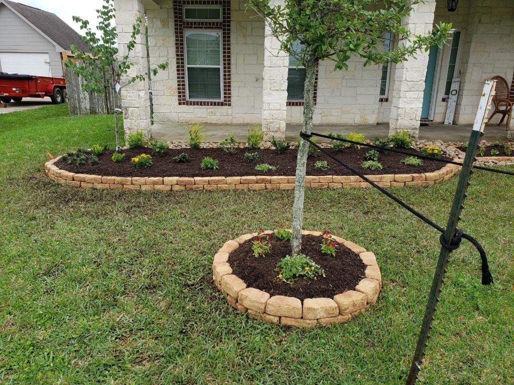 Front yard with brick-bordered flowerbeds, a young tree, and a house with beige siding.