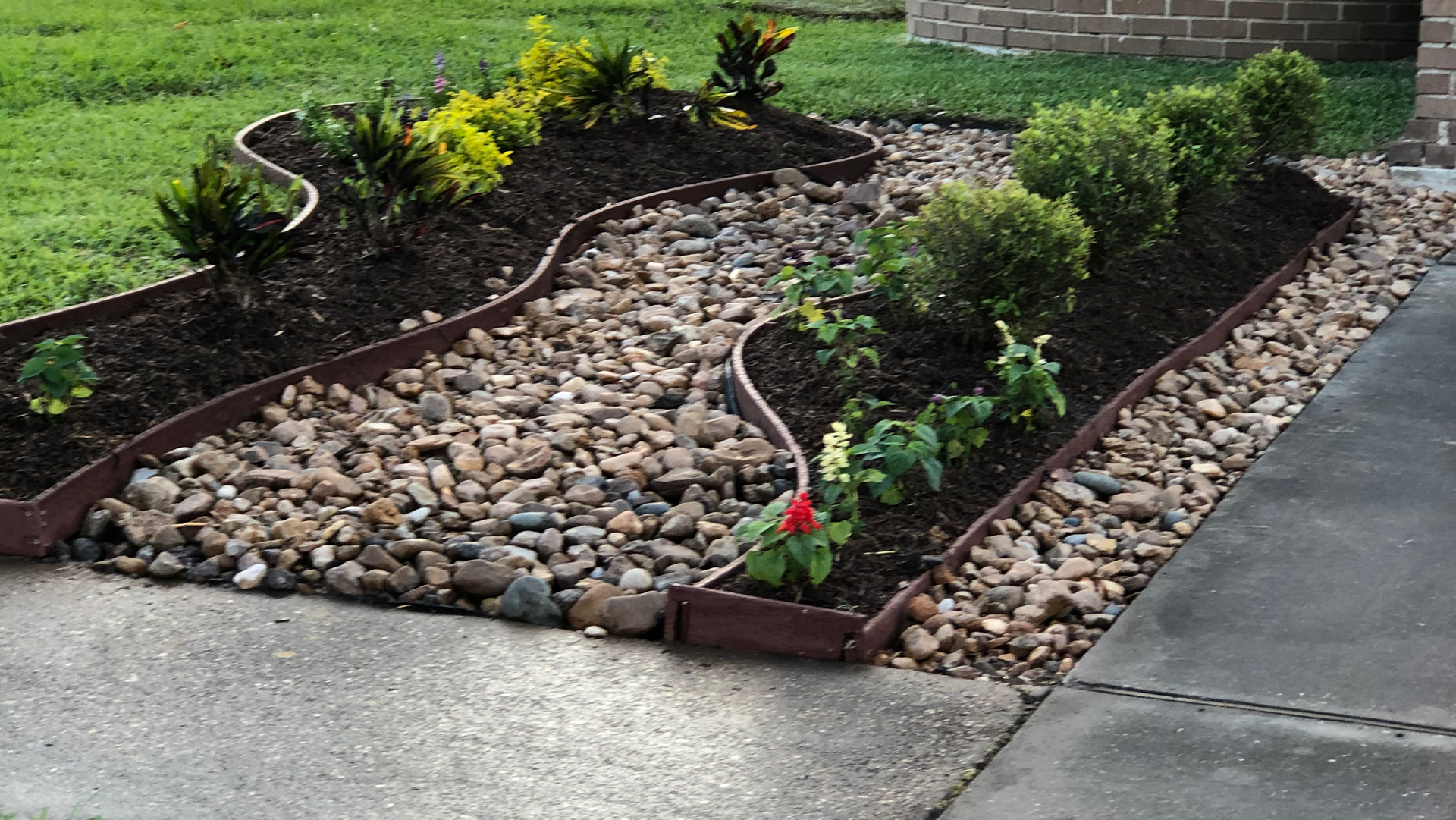 Landscape bed with dark soil, plants, brown rocks, and a brown border next to a sidewalk and grass.
