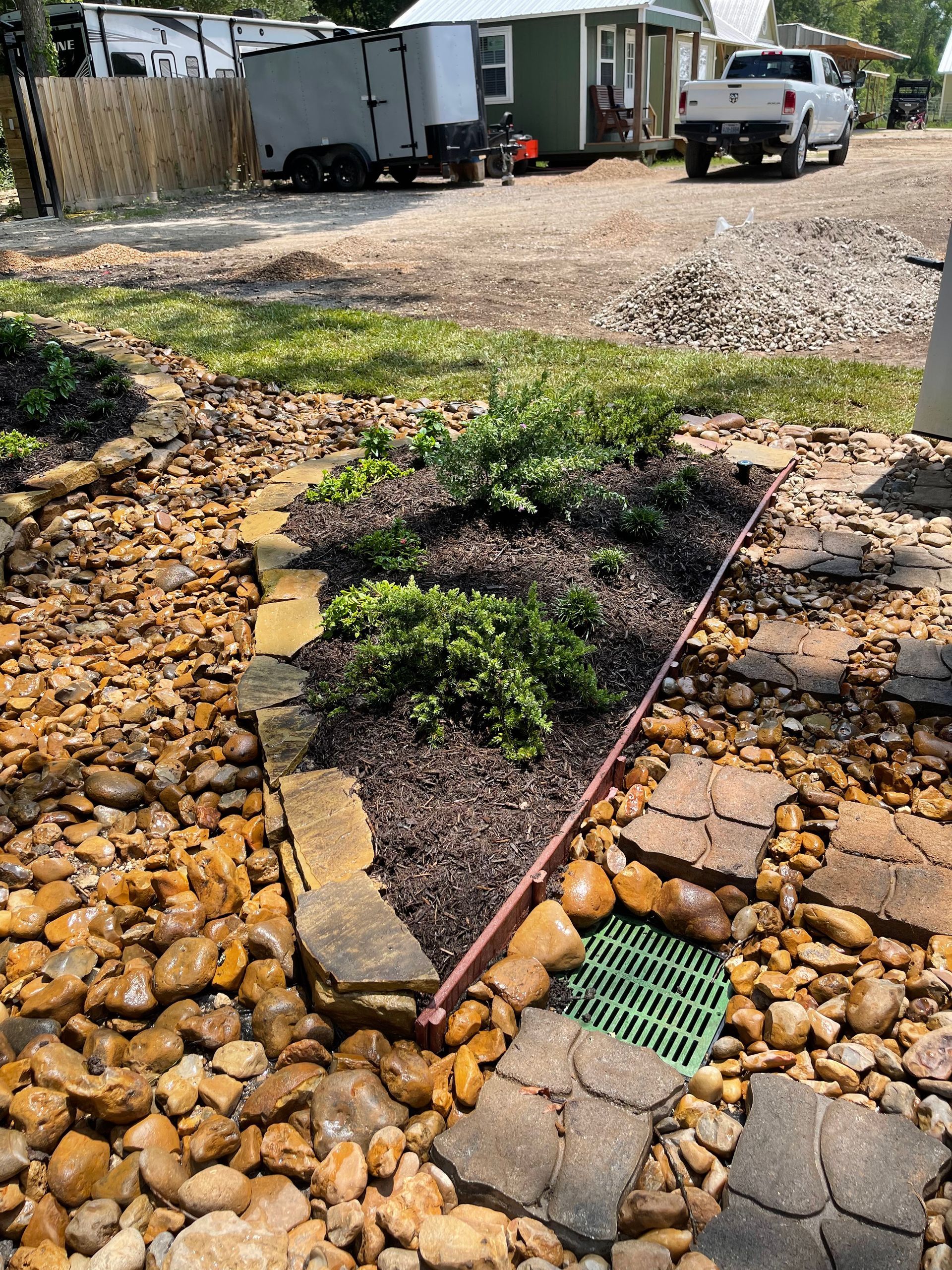 A garden bed with landscaping stones and plants, a drainage grate, and a driveway with vehicles.