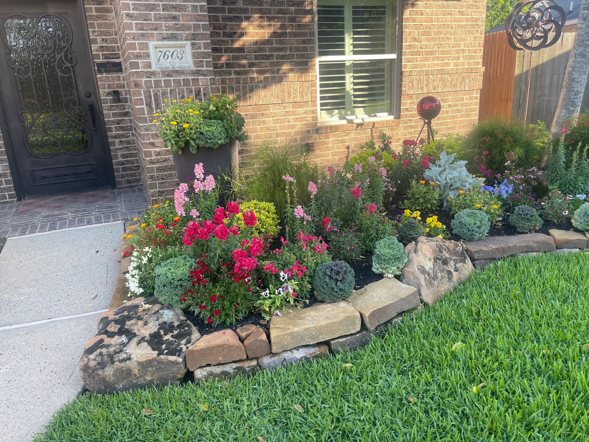 Colorful flower bed with brick edging and lush greenery in front of a brick house.