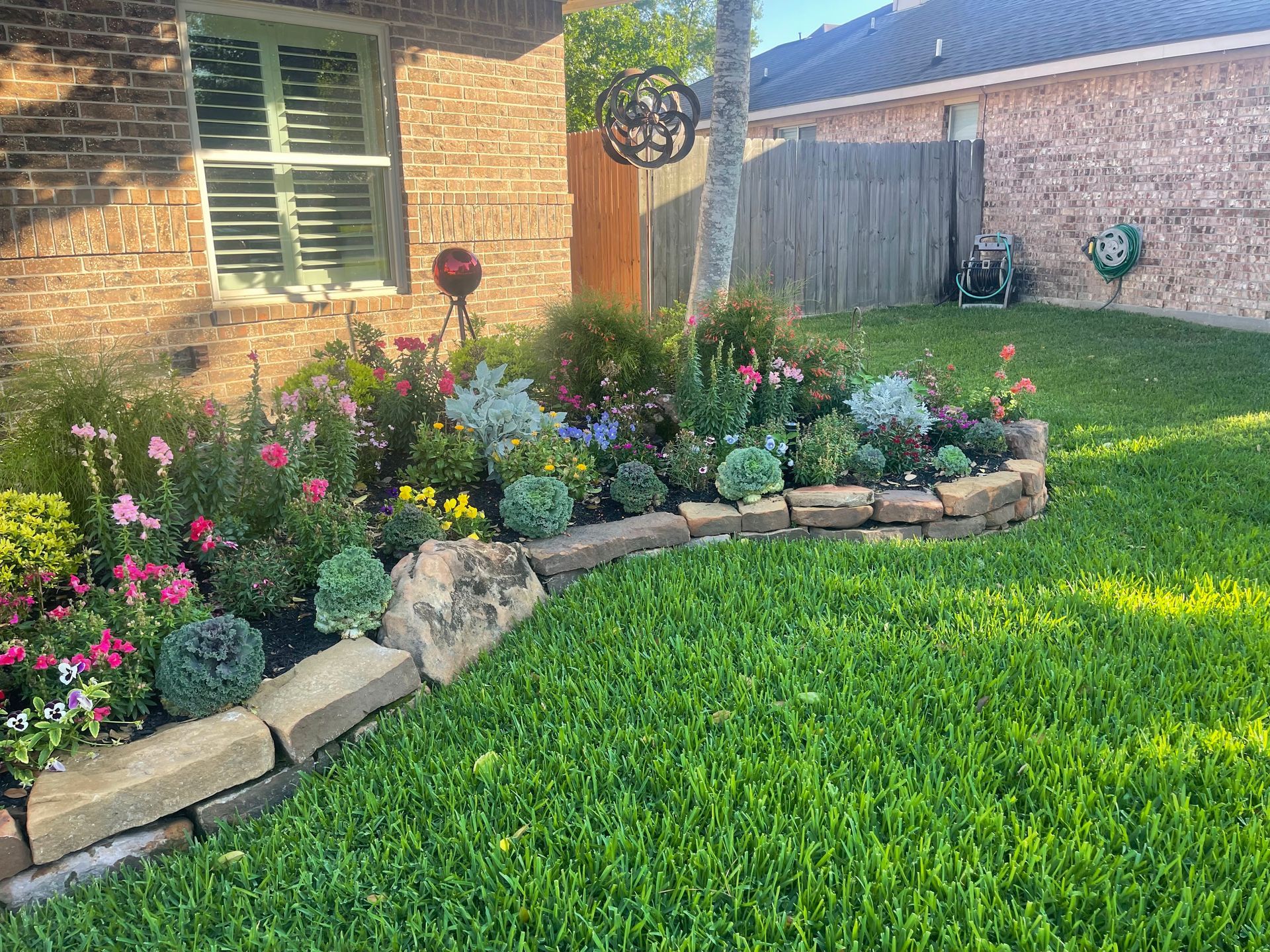 A vibrant flower bed edged with stone, filled with colorful plants, next to a brick house with green lawn.