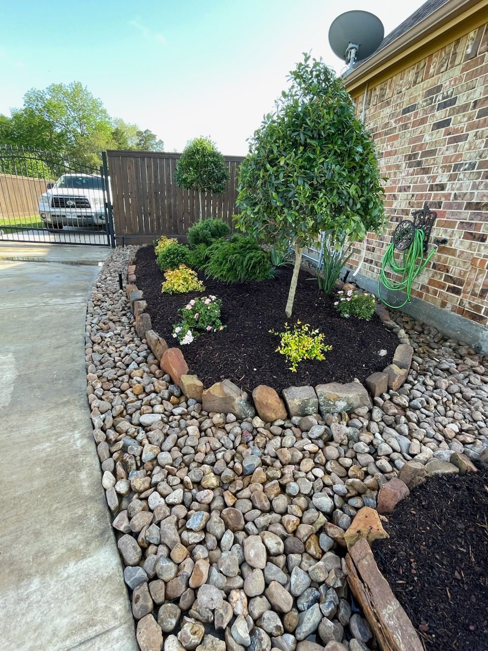 Landscaped garden bed with rocks, mulch, and plants next to a house and walkway.